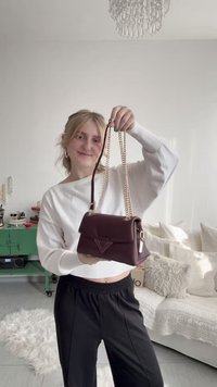 Mlaka žena u bijelom džemperu i crnim hlačama drži tamnoljubičastu kožnu torbu s lancem zlatne boje u svijetloj dnevnoj sobi.