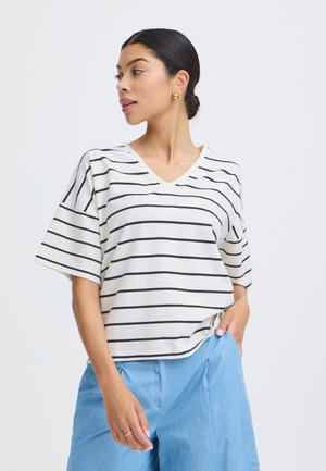 Kobieta w białej koszulce z krótkim rękawem i dekoltem w serek z czarnymi poziomymi paskami oraz jasnoniebieskich spodniach, stojąca z jedną ręką w kieszeni.