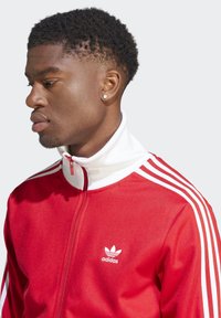 Red tracksuit jacket with white collar and zipper. Features three white stripes on sleeves and embroidered Adidas logo on chest. Smooth texture.