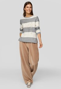 Langenärmliges Oberteil mit grauen und weißen Streifen und Schriftzug, kombiniert mit weiten beige Cordhosen. Model steht vor neutralem Hintergrund.