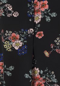 Blouse en tissu noir avec un motif floral comprenant des fleurs roses, bleues et jaunes. Conçue avec des boutons sur le devant et une texture lisse.