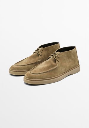 Brown suede desert boots with a rounded toe, stitched detailing, and minimal lacing. Rubber sole with a slightly raised profile.