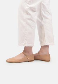 Pantofi Mary Jane bej din piele lacuită, cu curele la glezne, purtați cu pantaloni albi scurți, pe un fundal alb simplu.