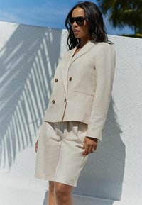 Lichtbeige dubbelzijdige blazer met gouden knopen en bijpassende op maat gemaakte shorts. Glad materiaal, relaxte pasvorm, minimalistisch ontwerp, gedragen met een zonnebril.