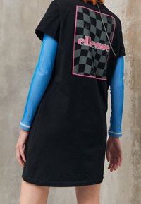 T-shirt noir avec un design à damier et des accents roses, portant le texte "ellesse". Manches longues bleuies côtelées sous le t-shirt.