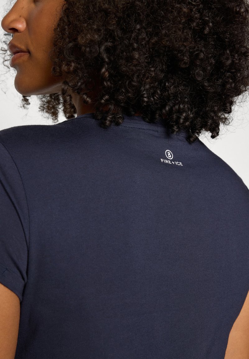 Frau mit lockigem Haar, die ein marineblaues Shirt trägt, auf dessen Rückseite sich nahe dem Ausschnitt ein kleines "FIRE + ICE"-Logo befindet.