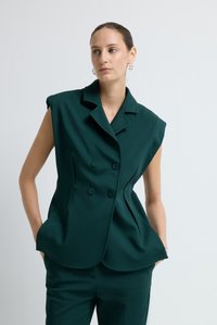 Žena s vlasy staženými dozadu, oblečená v tmavě zelené bezrukávové dvouřadové saku a odpovídajících kalhotách, stojící proti jednoduchému světlému pozadí.