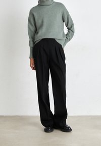 Grüner, gemütlicher Rollkragenpullover kombiniert mit schwarzen, hoch sitzenden, plissierten Hosen. Das Model trägt schwarze, runde Schuhe. Einfacher Hintergrund.