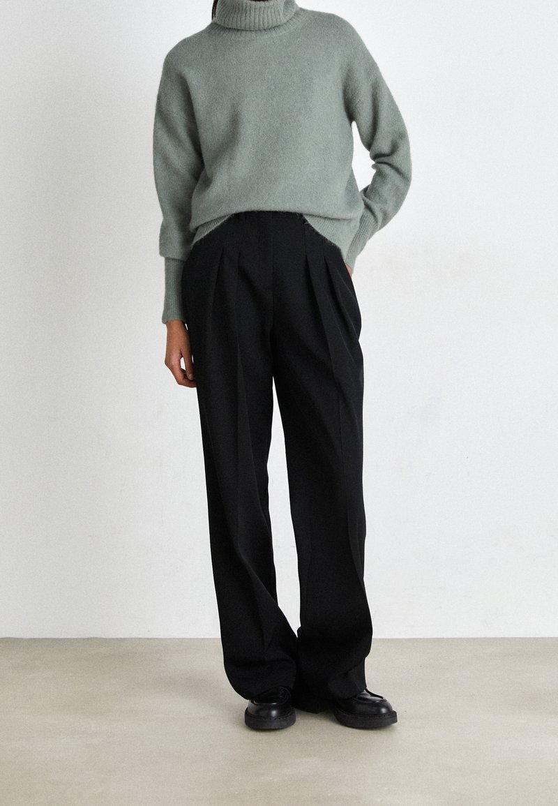Grüner, gemütlicher Rollkragenpullover kombiniert mit schwarzen, hoch sitzenden, plissierten Hosen. Das Model trägt schwarze, runde Schuhe. Einfacher Hintergrund.