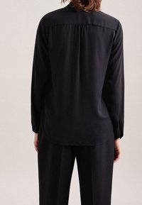 Blusa nera a maniche lunghe con una texture liscia, dettaglio plissettato sul retro e spacchi laterali. Indossata con pantaloni neri coordinati.