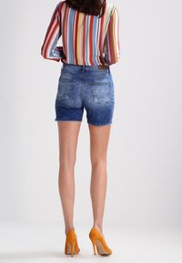 Nainen, joka on pukeutunut moniväriseen raidalliseen pitkähihaiseen paitaan, sinisiin farkkushortseihin ja oransseihin korkokenkiin, seisoo selin katsojaan.