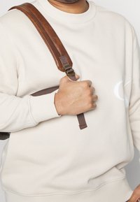 Beige Sweatshirts mit gerippten Bündchen, getragen von einer Person mit einem braunen Riemen einer Tasche vor einem neutralen Hintergrund. Das Logo ist auf dem Sweatshirt sichtbar.