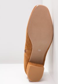 Semelle de chaussure en daim marron avec un petit talon carré, taille 37 indiquée, reposant contre une surface blanche et un mur.