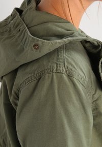 Olivgrüne Jacke mit Kapuze, aus strukturiertem Stoff, mit sichtbaren Nähten und Druckknopfdetails am Kragen.