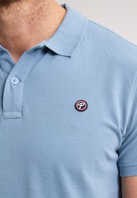 Lichtblauw poloshirt van gestructureerde stof, met een kraag met twee knopen en een rond logo-opdruk op de linkerkant van de borst.