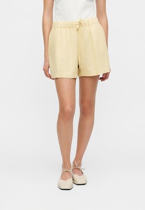 Vrouw draagt beige short met trekkoord, witte top en crèmekleurige ballerina's met enkelbandjes, staand tegen een effen achtergrond.