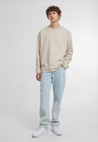 Beige Sweatshirt mit langen Ärmeln und dezentem Logo. Kombiniert mit hellblauen geraden Jeans und weißen Sneakers. Glatter Stoff, lockere Passform.