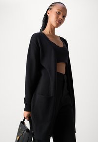 Cardigan noir oversize en tissu doux, présentant des manches longues, un devant ouvert et deux poches avant. Associé à un crop top noir.