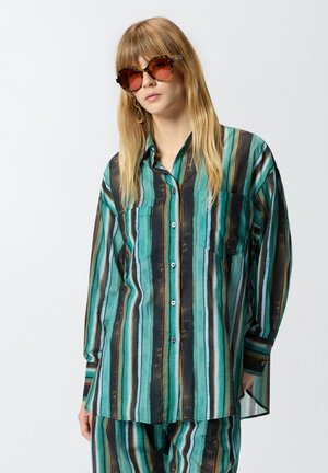 Personne aux longs cheveux blonds portant de grandes lunettes de soleil écaille de tortue et une chemise à rayures vertes, noires et marron boutonnée avec un pantalon assorti.