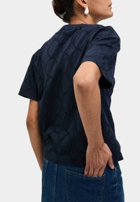 Námorně modrý textured top s krátkými rukávy, disponující vyšitým kulatým vzorem. V kombinaci s denimem, zobrazující zadní pohled a uvolněný střih.