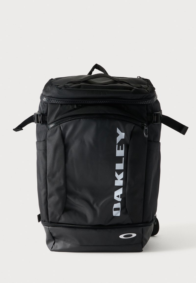 Schwarzer rechteckiger Oakley-Rucksack mit Tragegriff oben, verstellbaren seitlichen Riemen, Frontreißverschlusstasche und weißem Logo auf der Vorderseite.