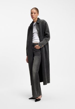 Vrouw die een lange donkere krijtstreeppak en bijpassende broek draagt over een wit overhemd, staand met één hand in de zak en zwarte puntige schoenen.