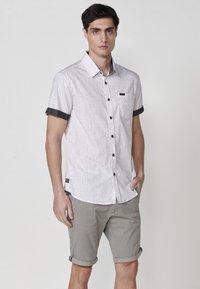 Camisa de botón de manga corta color gris claro con tejido texturizado, botones oscuros como acento y mangas enrolladas en contraste; combinada con pantalones cortos beige.