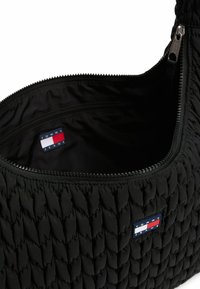 Borsa a spalla nera trapuntata con chiusura a zip. Presenta una superficie texture e un'etichetta interna con il logo di Tommy Jeans. Design compatto.