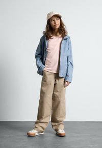 Hellblau gestreifte Jeansjacke, blassrosa T-Shirt, beigefarbene Weitbein-Hosen und beigefarbene Sneakers. Braune Mütze mit dezenten Mustern.