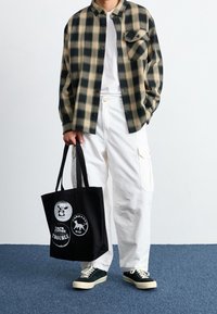 Chemise en flanelle à carreaux beige et noir, t-shirt blanc, pantalon cargo blanc et sac fourre-tout noir avec impressions graphiques. Baskets vertes.