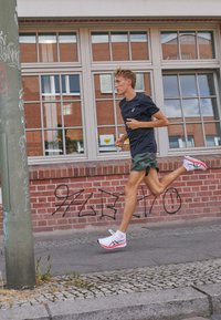 Löparskor med vit överdel, röda detaljer och svart logotyp. Löparen har på sig en mörk t-shirt och gröna shorts medan han joggar längs med en tegelvägg.