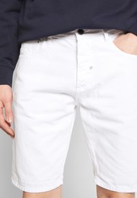 Pantaloni corti bianchi in cotone con una finitura leggermente strutturata, dotati di tasche anteriori, chiusura con bottone e cuciture lungo l'orlo.