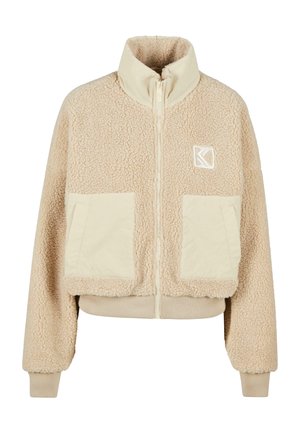 Beige fleece-takki, jossa on korkea kaulus, vetskari, kaksi etutaskua ja ribattu hihansuu. Vasemmassa rinnassa on logo.