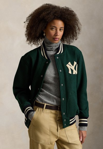Ciemnozielona kurtka varsity z kremowym logo NY, w paski ściągacze przy mankietach i zatrzaskami. Narzucona na szary golf i beżowe spodnie.