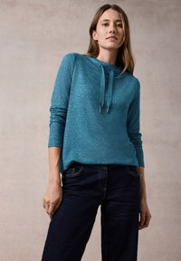 Teal Pullover mit strukturierter Oberfläche, ausgestattet mit Kapuze und langen Ärmeln. Kombiniert mit dunklen Jeans und sichtbaren Nähten.