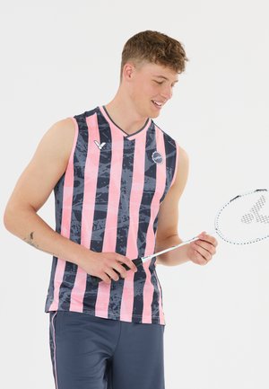 Ung mand i pink og sort stribet sportstøj uden ærmer, der holder en badmintonketcher, kigger ned og smiler.