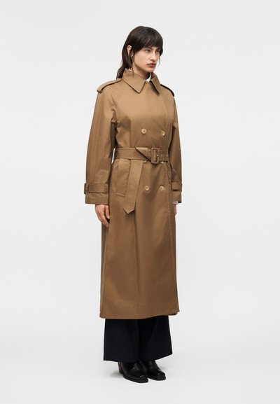 Γυναίκα που φορά μακρύ καμηλό διπλόπατο παλτό με ζώνη, στέκεται με μαύρο παντελόνι φαρδιάς γραμμής και μαύρα μπότες με τακούνι σε απλό φόντο.