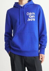 Blauwe katoenen hoodie met een voorzak, trekkoordcapuchon en een bedrukte logo met de tekst "Calvin Klein Jeans." Geribbelde manchetten en zoom.