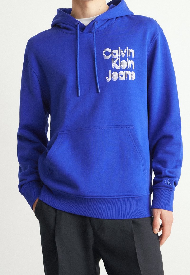 Blauwe katoenen hoodie met een voorzak, trekkoordcapuchon en een bedrukte logo met de tekst "Calvin Klein Jeans." Geribbelde manchetten en zoom.