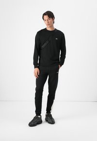 Black sweatshirt and joggers with a fitted design. Notable green logo on the chest and side stripes on pants. Black sneakers complete the look.