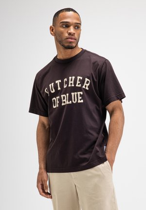 Man die een donkerbruine T-shirt met de tekst "BUTCHER OF BLUE" en beige broek draagt, kijkt opzij met één hand in de zak.