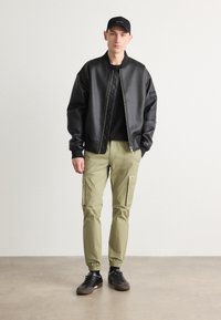 Zwarte neplederen bomberjack met ribbelmanchetten en -zoom, gecombineerd met lichtgroene cargobroek met zijzakken en elastische manchetten. Zwarte sneakers.