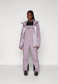 Moteris, dėvinti levandų spalvos slidinėjimo striukę ir kombinezoną, su juodais slidinėjimo batais, stovinti prieš paprastą šviesiai pilką foną.