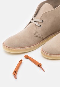 Botas de tobillo de ante beige con costuras en tono marrón y suelas de goma crema. Dos cordones naranjas están colocados cerca.