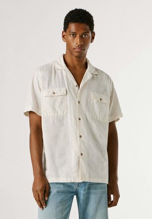 Giovane uomo che indossa una camicia crema a maniche corte con bottoni e due tasche sul petto, abbinata a jeans azzurri, in piedi davanti a uno sfondo semplice.