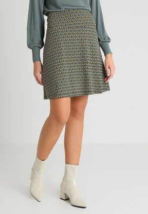 Ein bedruckter Rock mit geometrischen Formen in Grün, Gelb und Schwarz, kombiniert mit cremefarbenen Stiefeletten, vor einem neutralen Hintergrund.