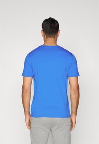 Μπλε κοντομάνικη μπλούζα με στρογγυλή λαιμόκοψη, φτιαγμένη από μαλακό ύφασμα, εμφανίζεται από πίσω. Φορεμένη με γκρίζο παντελόνι.