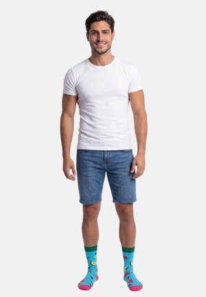 Smilende mand stående iført en hvid t-shirt, blå denimshorts og blå sokker med farverige frugtmønstre.