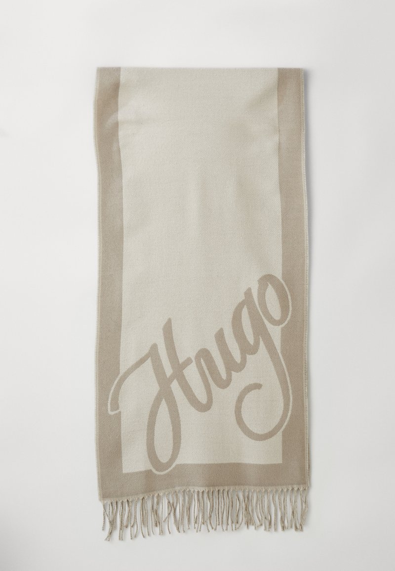 Foulard rectangulaire beige et crème avec des bords frangés et "Hugo" en grandes lettres cursives sur un côté.