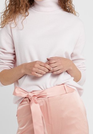 Femme portant un pull à col roulé rose clair et un pantalon rose en soie avec une ceinture nouée, les mains légèrement jointes au niveau de la taille.